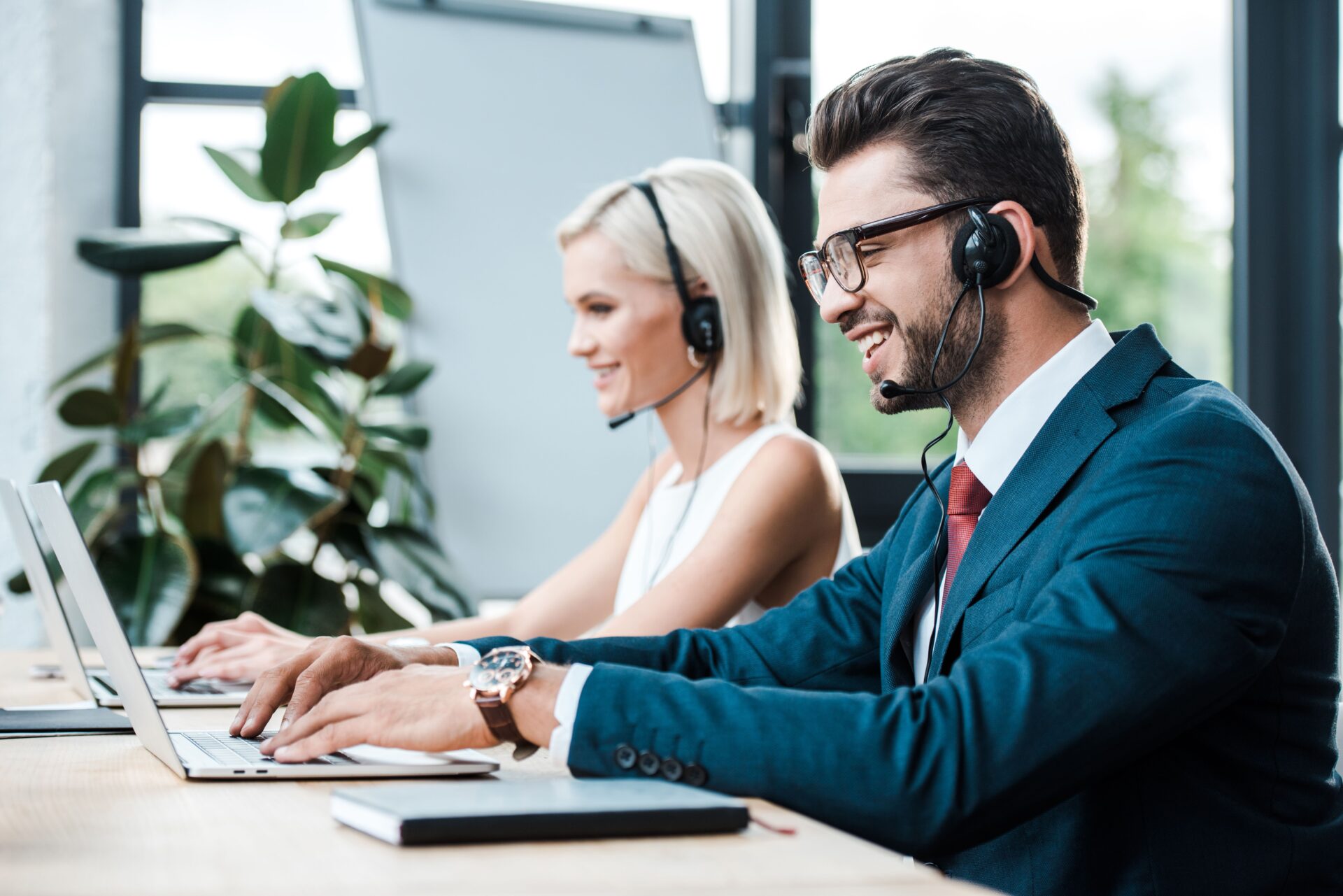 selective focus of happy operator in headset typing on laptop near blonde colleague in headset selective focus of happy operator in headset typing on laptop near blonde colleague in headset